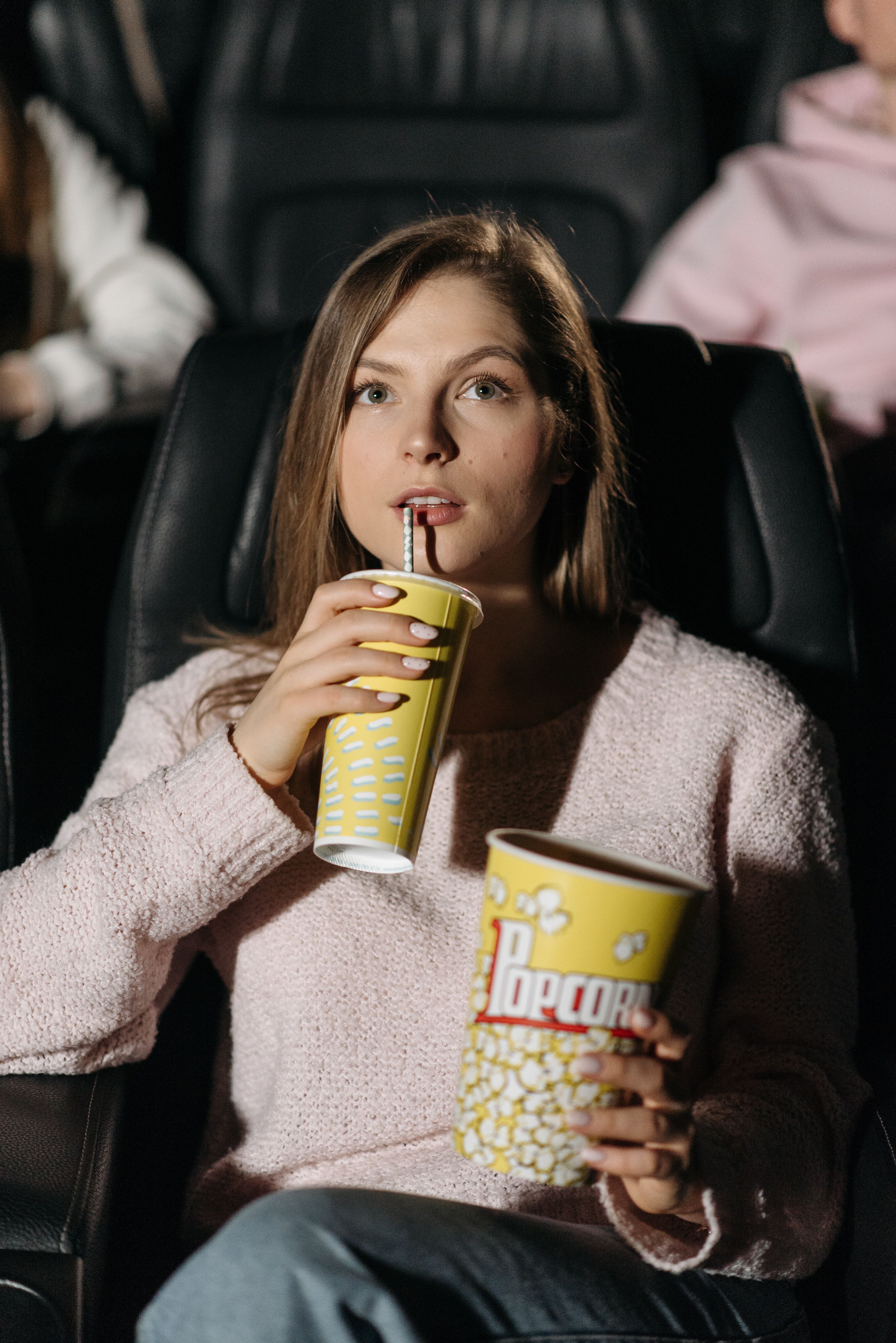 A woman sitting in a cinema drinking a soft drink and holding a bucket of popcorn