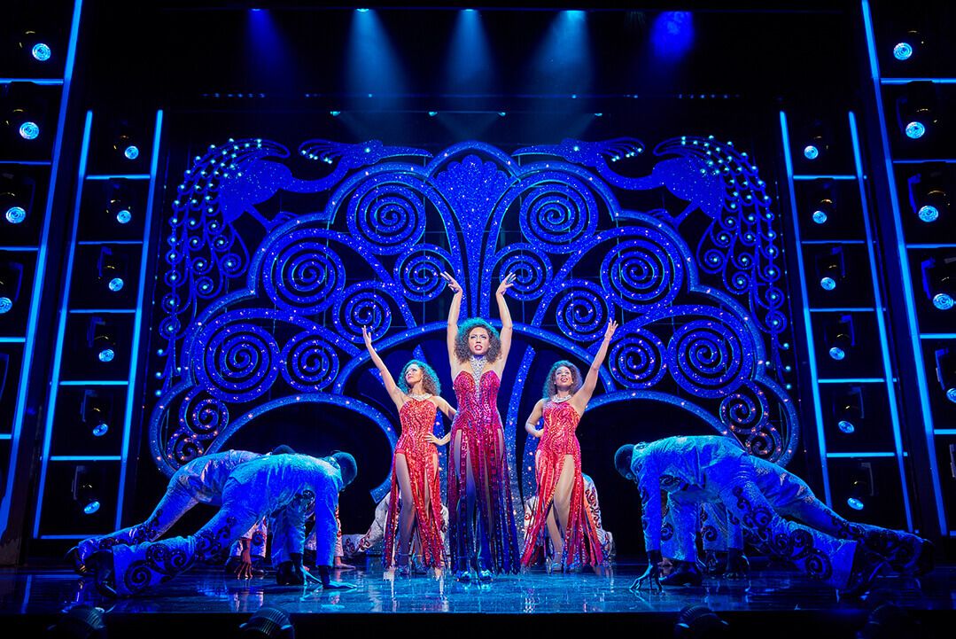 Three performers in red sequinned dresses on stage during a Dreamgirls performance, lit in blue with an ornate backdrop