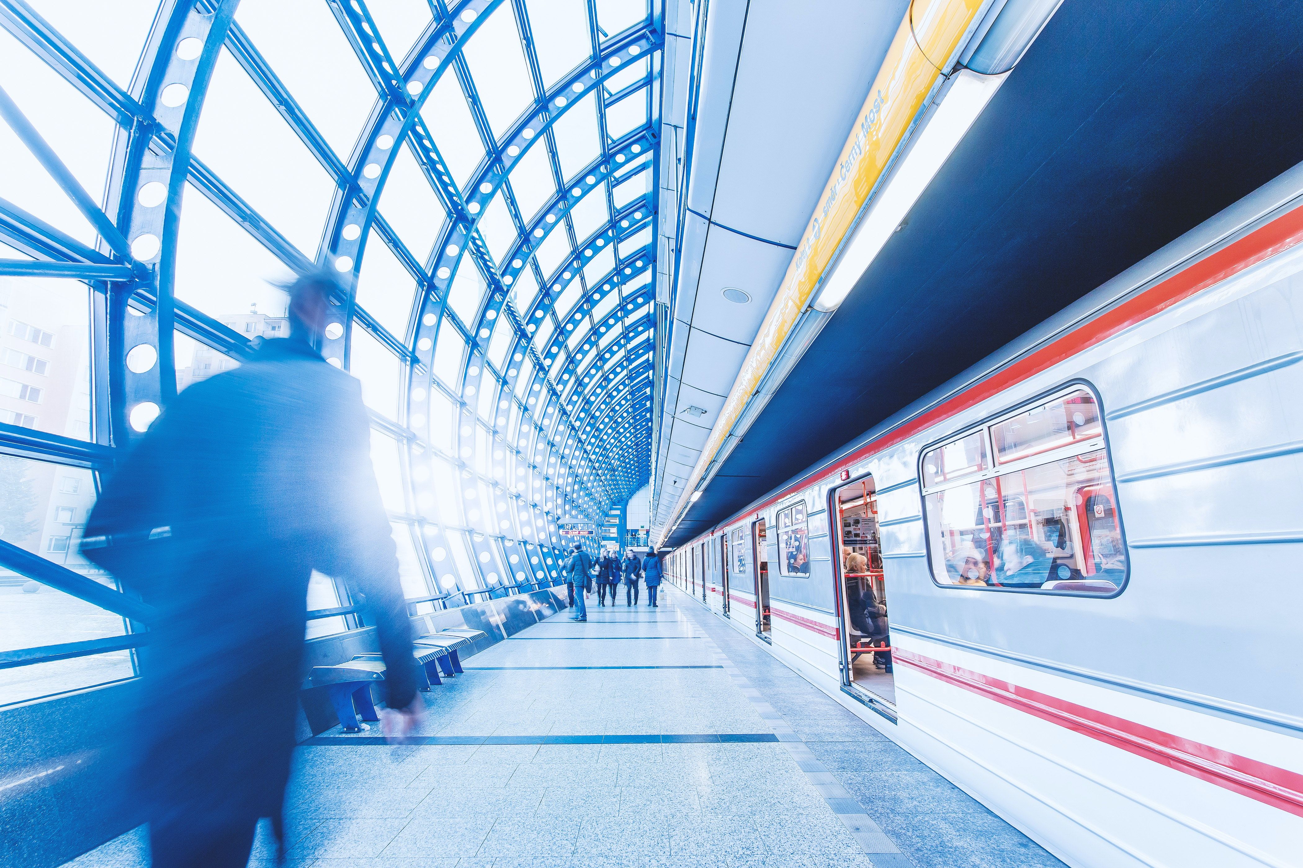 Blurred commuters walking along a bright, curved-glass train platform as a train waits with its doors open