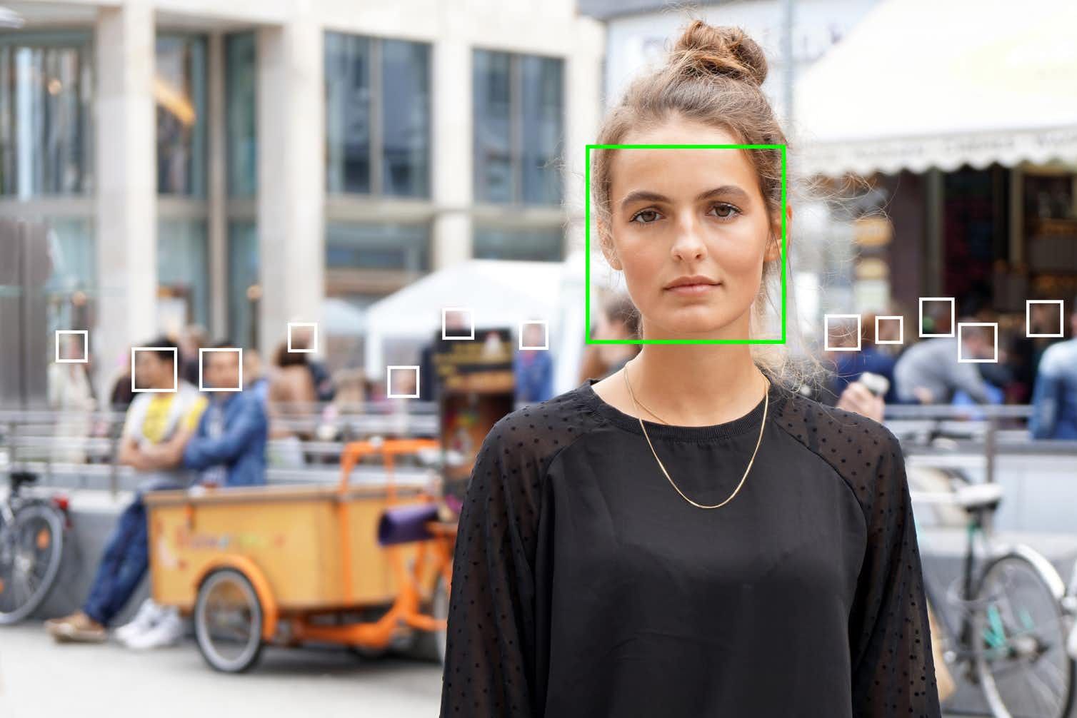 Facial recognition software analysing a woman's face in a street scene, with detection boxes drawn around faces in the crowd